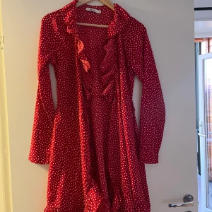 Röd prickig klänning från NA-KD - Säljer en röd klänning med vita prickar från NA-KD. Klänningen har långa ärmar och volangdetaljer längs kragen och fram. Perfekt för en färgglad look!