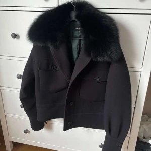 Svart kappa med pälsdetalj från Meotine - Elegant svart kappa från Meotine med en lyxig pälsdetalj vid kragen. Jackan har knappar framtill och två fickor med lock. Perfekt för att hålla stilen i vår väder. Pris går att diskuteras☺️