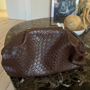 Brun clutch i syntet - Snygg brun clutch med ett elegant rutmönster. Väskan är tillverkad i syntetmaterial och har en rymlig öppning med guldfärgade detaljer. Perfekt för att bära med sig det nödvändigaste med stil.