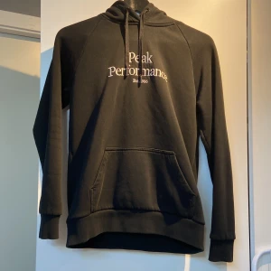 Svart hoodie från Peak Performance - Säljer en svart hoodie från Peak Performance med vit text på bröstet. Tröjan har en klassisk känguruficka och justerbar huva med dragsko. Perfekt för en avslappnad stil.