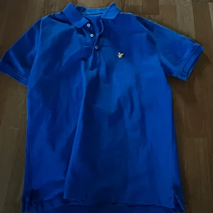 Blå pikétröja med broderad logga - Snygg blå pikétröja med en broderad logga på bröstet. Tröjan har en klassisk krage och knappar framtill. Perfekt för en avslappnad stil.