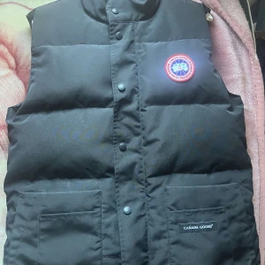Svart dunväst från Canada Goose - Säljer en svart dunväst från Canada Goose med knappar och två praktiska fickor framtill. Västen har en klassisk design med en rund logotyp på bröstet. Passar året runt. Bilderna ser lite dåliga ut i kvalite men de är pga min söndriga kamera däremot så är den nästan aldrig använd och i topp skick  