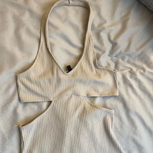 Beige ribbad klänning - Snygg beige ribbad klänning med en djup v-ringning och halterneck-design. Klänningen har en tight passform och är ärmlös, vilket ger en stilren look. Perfekt för en somrig dag eller kväll.