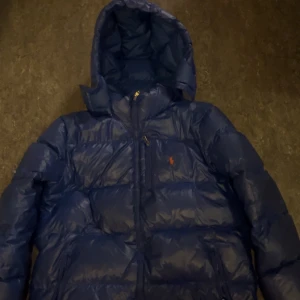 Blå dunjacka med huva - Säljer en blå dunjacka från Polo Ralph Lauren. Jackan har en praktisk huva och dragkedja framtill. Perfekt för kalla dagar.