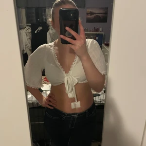 Vit omlottopp - Säljer en söt vit omlottopp med knytning framtill och spetsdetaljer längs kanterna. Toppen har korta ärmar och är croppad, vilket ger en trendig look. Perfekt för varma dagar eller sommarens fester! Aldrig används så den är i nyskick