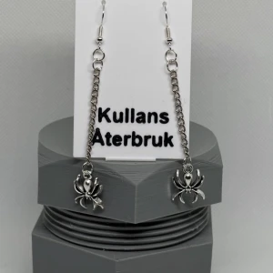 Silverfärgade örhängen med spindelmotiv - Unika silverfärgade örhängen från Kullans Återbruk med ett coolt spindelmotiv. Örhängena hänger i en kedja och ger en edgy look. Perfekta för att ge din outfit en extra touch!
