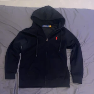  Polo Ralph Lauren hoodie - Säljer en stilren svart hoodie från Polo Ralph Lauren med dragkedja och en röd broderad logotyp på bröstet. Tillverkad i 100% bomull för en bekväm känsla. Perfekt för en avslappnad look.