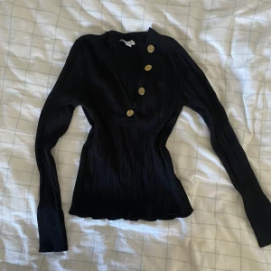 Svart tröja med gulddetaljer  - Svart ribbad tröja med guldknappar. V-ringad. Storlek M, men skulle säga mer en S.