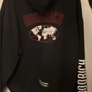 Svart hoodie från Hoodrich - Säljer en svart hoodie från Hoodrich med tryck av en världskarta och texten 'HOODRICH' i rött på ryggen. Framsidan har en stor ficka och flera logotyper. Perfekt för en avslappnad stil. Den är välanvänd med inga som helst skador. Köpt i en hoodrich butik i Stockholm.