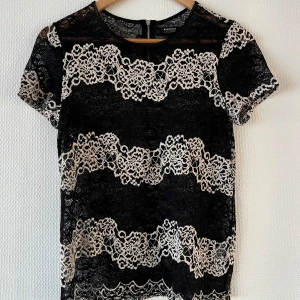 Svart spetsblus med korta ärmar - Elegant svart blus med vackra spetsdetaljer i vitt. Blusen har korta ärmar och en rund halsringning, perfekt för en stilren look. Den är tillverkad i ett lätt och luftigt material som ger en bekväm passform. Liknar intimissimis ikoniska spets🖤