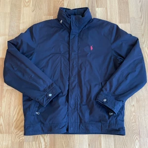 Mörkblå jacka från Polo Ralph Lauren - Säljer en snygg mörkblå jacka från Polo Ralph Lauren med en diskret röd logga på bröstet. Jackan har långa ärmar och stängs med dragkedja och knappar. Den är fodrad med fleece för extra värme och har en stilren design.