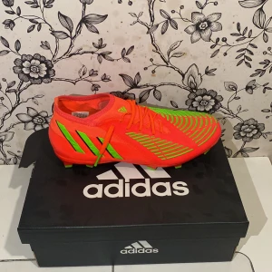 Adidas Predator fotbollsskor - Snygga Adidas Predator fotbollsskor i en iögonfallande röd och grön färgkombination. Skorna har snörning och är designade för optimal prestanda på planen. Perfekta för den som vill sticka ut och dominera spelet.