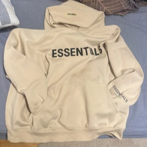 Beige hoodie från Essentials - Säljer en stilren beige hoodie från Essentials by Fear of God. Den har en stor logotyp på framsidan och en mindre på ärmen. Perfekt för en avslappnad look med en känguruficka framtill och ribbade muddar.