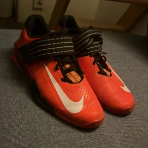 Nike weightlifting  - Snygga röda Nike träningsskor med svart och vit detaljering. Skorna har snörning och ett brett band över foten för extra stöd. Perfekta för gymmet eller annan träning.