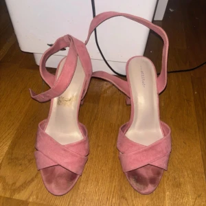Rosa sandaler med klack - Säljer ett par eleganta rosa sandaler med klack och korsade remmar. De har en justerbar rem runt ankeln för bättre passform. Perfekta för sommarens alla tillfällen.