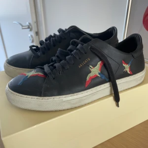 Svarta sneakers från Axel Arigato - Snygga svarta sneakers från Axel Arigato med broderade fåglar i rött, blått och grönt på sidorna. Skorna har en vit sula och svarta snören. Perfekta för en stilren look.
