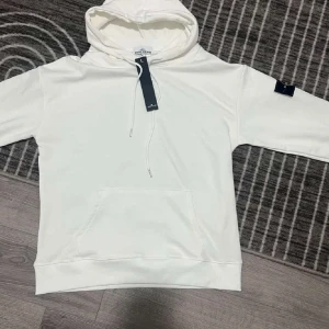 Vit hoodie från Stone Island - Säljer en stilren vit hoodie från Stone Island med klassisk logga på ärmen. Hoodien har en känguruficka och justerbar huva med snören. Perfekt för en avslappnad stil.