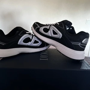 Dior 30 - Säljer ett par snygga Dior sneakers i svart och vitt med tydliga logodetaljer och cool design. Skorna har snörning, rund tå och en tjock, platt sula som ger en sportig vibe. Perfekta för dig som vill sticka ut med exklusiva sneakers. B30
