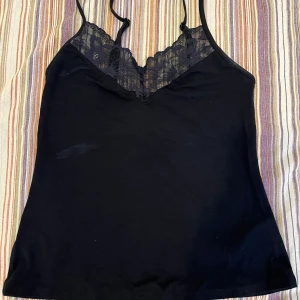 Svart linnetopp med spetsdetaljer - Säljer en svart linnetopp med tunna axelband och v-ringning dekorerad med blommig spets framtill. Fläck på framsida som förmodligen är deo och försvinner i tvätten, kan prova tvätta innan köp och höra av mig.