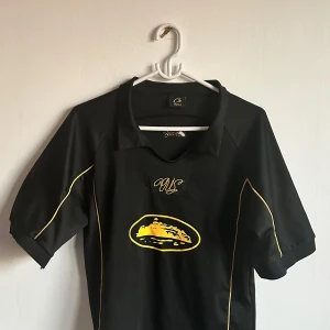 Svart fotbollströja med gula detaljer från Corteiz  - Säljer en svart fotbollströja från Corteiz med gula linjer längs sidorna och ett gult tryck på bröstet. Tröjan är tillverkad i 100% polyester och har korta ärmar. Perfekt för träning eller match!