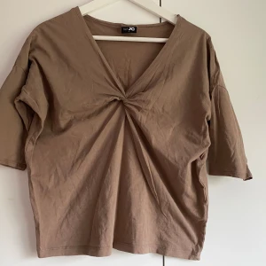 Beige v-ringad topp från JC - Säljer en beige topp från JC med v-ringning och twistad detalj framtill. Toppen har korta ärmar och är tillverkad i mjuk bomull. Perfekt för en avslappnad och stilren look.