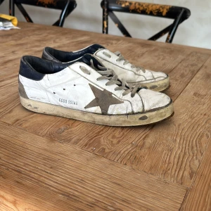 Golden Goose med grå stjärna - Säljer ett par vita sneakers från Golden Goose med grå stjärndetalj på sidan och svarta detaljer vid hälen. Skorna har snörning och platt sula, perfekt för en avslappnad stil och till sommaren. Dock behöver skorna lagas då dom är i dåligt skick där av priset. Vet inte häller storleken helt men jag har 42 och de passar mig perfekt 