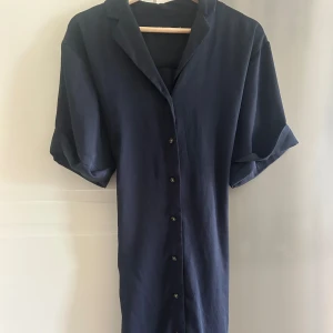 Marinblå skjortklänning med knappar - En stilren marinblå skjortklänning med korta ärmar och knappar längs hela framsidan. Klänningen har en klassisk krage och är tillverkad i ett mjukt, lätt material. Perfekt för en avslappnad men snygg look.