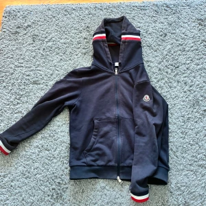 Mörkblå hoodie från Moncler med dragkedja - Säljer en mörkblå hoodie från Moncler med hel dragkedja och huva. Tröjan har vita och röda ränder på muddar och huva samt Moncler-logga på bröstet. Saknar dock snören.