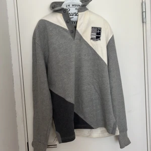 Grå och vit långärmad pikétröja från InvidiaUomo - Säljer en långärmad pikétröja från InvidiaUomo i storlek Large. Tröjan har blockmönster i olika nyanser av grått och vitt, knappslå vid halsen och en broderad patch på bröstet. Skön och stilren modell med klassisk krage.