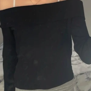 Svart ribbad offshoulder-top från H&M - Säljer en svart ribbad offshoulder-top från H&M. Toppen har en bred kant upptill som sitter snyggt över axlarna och är tillverkad i ett stretchigt material. Perfekt att styla med jeans eller kjol.