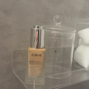 CAIA Dewy Drops Serum Foundation - Flytande foundation från CAIA i nyansen Dewy Drops. Kommer i en glasflaska med silvrig pump och lock. Ger en naturlig, återfuktande finish och är enkel att applicera för en fräsch look.