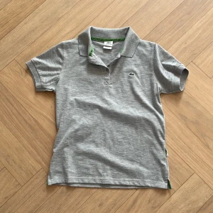 Grå pikétröja från Lacoste - Säljer en klassisk grå pikétröja från Lacoste med liten broderad logga på bröstet. Tröjan har krage, korta ärmar och knäppning framtill. Perfekt för en stilren och sportig look, Skriv till mig vid minsta tanke! Storlek XL i barn passar barn som är 12år eller 11år