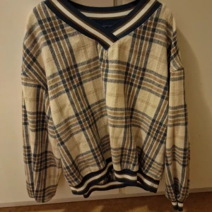 Beige och blå rutig tröja med v-ringning - Säljer en oversized tröja med v-ringad krage i beige och blått rutigt mönster. Tröjan har långa ärmar och ribbade muddar vid ärmslut och nederkant. Perfekt för dig som gillar retrostil!