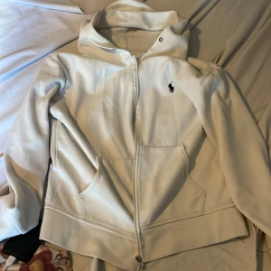 Vit hoodiejacka från Polo Ralph Lauren - Vit hoodiejacka från Polo Ralph Lauren med dragkedja framtill och två fickor. Klassisk logga broderad på bröstet. Perfekt för en avslappnad stil och enkel att matcha med det mesta.