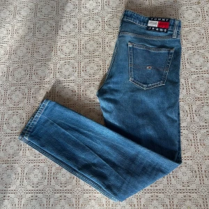 Blå jeans från Tommy Jeans - Säljer ett par klassiska blå jeans från Tommy Jeans med normal passform. Byxorna har fem fickor, raka ben och ikonisk Tommy Jeans-logga på bakfickan och linningen. Perfekta för dig som gillar en tidlös och enkel stil. Storlek 30 32. 