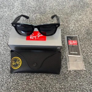 Svarta Wayfarer solglasögon från Ray-Ban - Klassiska svarta Wayfarer solglasögon från Ray-Ban med polaroid-linser. Bågarna är i blank svart plast och har Ray-Ban-logga på både glas och skalmar. Säljes med originalfodral och putsduk.