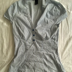 Figursydd blus - Säljer en blåvit randig blus med v-ringning och korta ärmar. Klänningen har knappar framtill och markerad midja för en smickrande passform. Perfekt för varma dagar och enkel att styla. I fint skick. Står storlek 38 men passar xs-s 💕