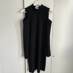 Svart långärmad klänning med öppna axlar från Vero Moda - Säljer en svart långärmad klänning från Vero Moda med en lite högre krage och snygga cut out-detaljer vid axlarna. Klänningen har en rak passform och är tillverkad i ett mjukt, stretchigt material. Perfekt för dig som gillar minimalistisk stil. Saknar knappar på baksidan.