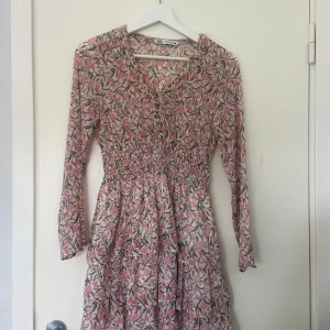Blommig kortklänning från Zara - Söt kortklänning från Zara i blommigt mönster med rosa och gröna toner. Klänningen har v-ringning, smockad midja och långa ärmar. Perfekt för vår och sommar, lätt och luftig i materialet.