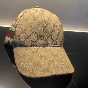 Beige keps från Gucci med GG-mönster - Säljer en beige keps från Gucci med klassiskt GG-monogram över hela kepsen och grönt/rött band på sidan. Kepsen har böjd skärm och justerbar passform. Perfekt för dig som gillar lyxiga accessoarer.