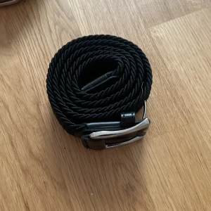 Svart flätat bälte med silverfärgat spänne - Säljer ett svart flätat bälte med silverfärgat metallspänne. Bältet har detaljer i svart skinn vid spännet och änden. Passar perfekt till jeans eller chinos och ger en stilren look.