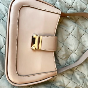 Beige axelväska med gulddetalj - Säljer en stilren beige axelväska med snygg guldfärgad spänndetalj framtill. Väskan har en mjukt rundad form och är tillverkad i ett slätt material. Perfekt för dig som gillar minimalistisk design och vill ha en väska som passar till många outfits.
