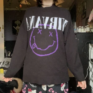 Svart Nirvana-tröja med lila smiley - Cool svart långärmad tröja med stort Nirvana-tryck och lila smiley på framsidan. Oversized passform och mjukt material, perfekt för dig som gillar bandtröjor och vill ha en avslappnad stil.