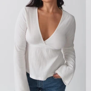 Vit omlottopp med v-ringning och knytband - Säljer en vit omlottopp med djup v-ringning och långa ärmar. Toppen har knytband i midjan som ger en fin passform och är tillverkad i ett lätt, strukturerat material. Perfekt att styla med jeans eller kjol.