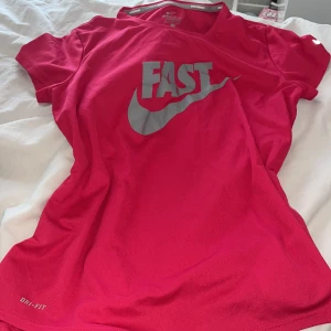 Rosa Dri-Fit t-shirt från Nike - Säljer en rosa Nike Dri-Fit t-shirt med silverfärgat 'FAST'-tryck och swoosh på bröstet. Perfekt för löpning eller träning, tillverkad i funktionsmaterial som andas. Klassisk passform med korta ärmar och rund hals.