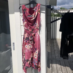 Blommig klänning från Billie & Blossom - Säljer en blommig klänning från Billie & Blossom i en vinröd nyans. Klänningen har omlottkänsla, knytband i midjan och dragkedja bak. Mönstret består av stora blommor i vitt, orange och blått. Perfekt för sommarens tillfällen! Storlek L men är liten i storleken!