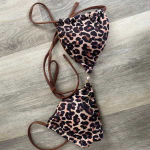 Leopardmönstrad bikini med knyt - Säljer en snygg trekantsbikini med leopardmönster och bruna knytband. Bikinin har gulddetalj framtill och är tillverkad i ett stretchigt syntetmaterial med elastan. Passar bra till en svart underdel, Storlek XS 