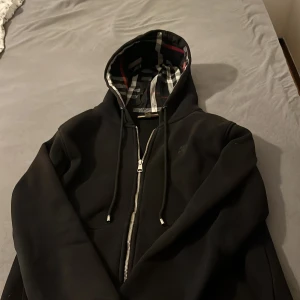 Svart hoodieZip från Burberry - Säljer en svart hoodiejacka från Burberry med klassiskt rutigt foder i svart, vitt och rött. Jackan har huva med dragsko och är helsvart på utsidan. Perfekt för dig som gillar stilrena plagg med lyxig känsla. Knappt använd då den är för liten för mig.