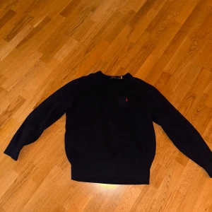 Mörkblå sweatshirt från Polo Ralph Lauren - Säljer en klassisk mörkblå sweatshirt från Polo Ralph Lauren med rund hals och den ikoniska röda loggan broderad på bröstet. Tröjan har långa ärmar och är tillverkad i mjuk bomull. Perfekt för en stilren och avslappnad look. Köpt för 2100kr. Pris går att diskutera.