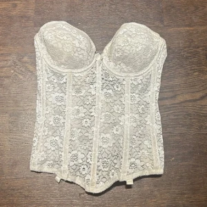 Vit spetskorsett - Säljer en vit korsett med vacker blommig spets och vadderade kupor. Toppen har en klassisk korsettform med markerade sömmar och hakknäppning bak. Perfekt för att skapa en feminin och elegant look.